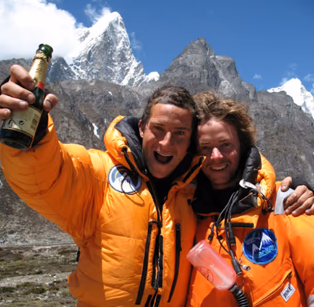 Two climbers in bright orange jackets celebrate with a bottle and smile in front of snowy mountain peaks under a blue sky.
