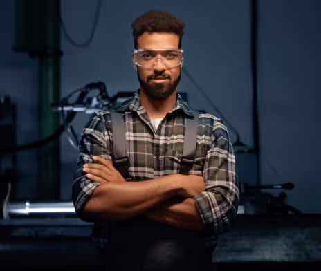 Man wearing safety glasses and a plaid shirt with folded arms in a workshop or industrial setting.