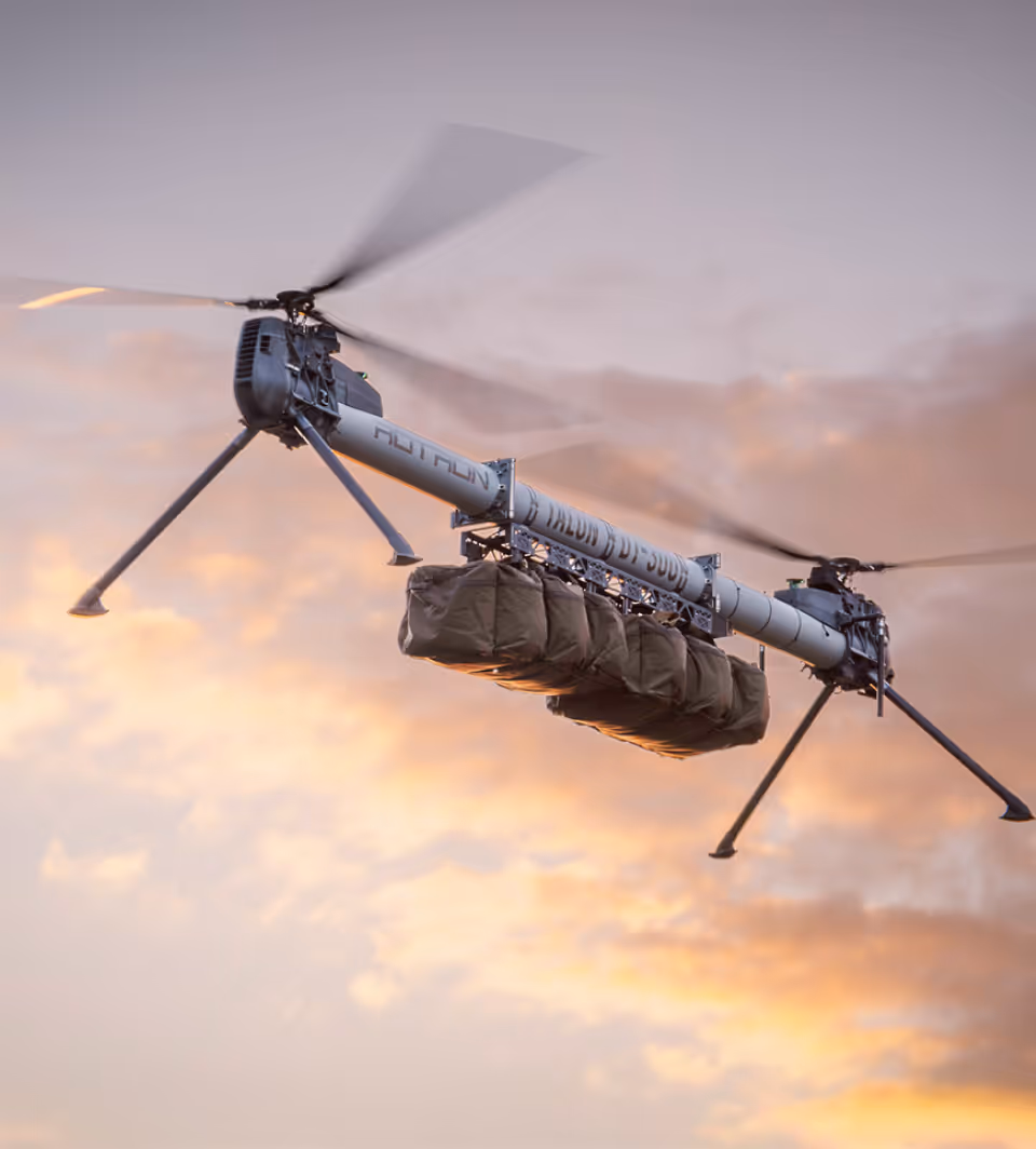 A long, twin-rotor drone carrying six large cargo bags, flying against a sunset sky.