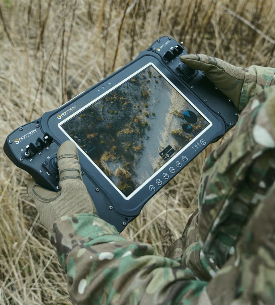 Soldier in camouflage gloves holding a Rotron remote control device displaying a landscape view with charts.