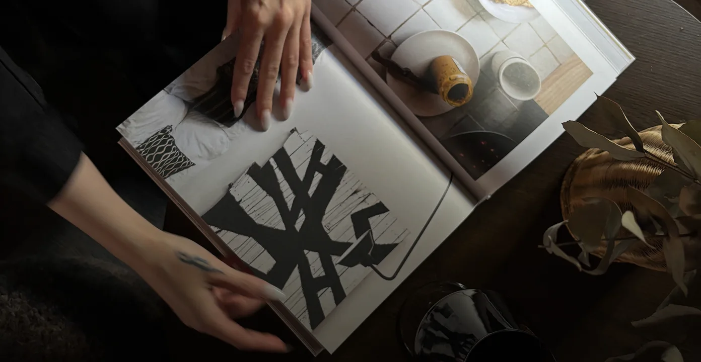 Person with nail art and a hand tattoo flipping through a magazine showing abstract black-and-white and kitchen images on a wooden table.