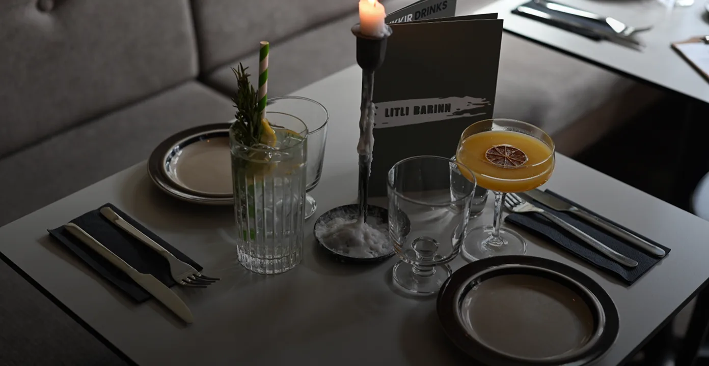 Table set for two with empty plates, utensils on black napkins, two drinks, a lit candle, and a menu labeled 'LITLI BARINN'.