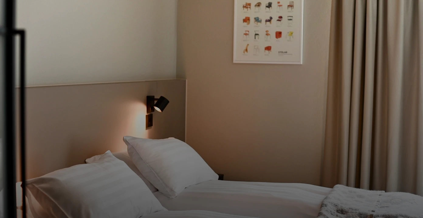 Minimalist bedroom with a neatly made bed, two white pillows, a wall-mounted reading lamp, a framed poster of chairs, and beige curtains.