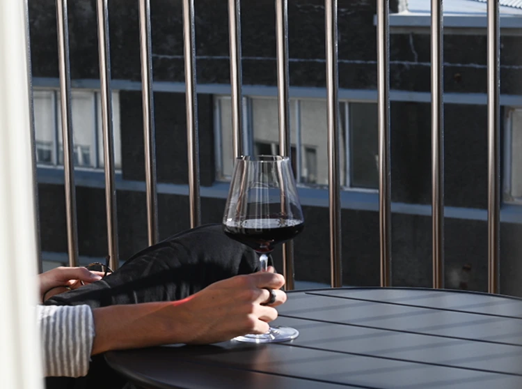 Person sitting on a balcony holding a glass of red wine next to a black table with vertical railing bars in the background.