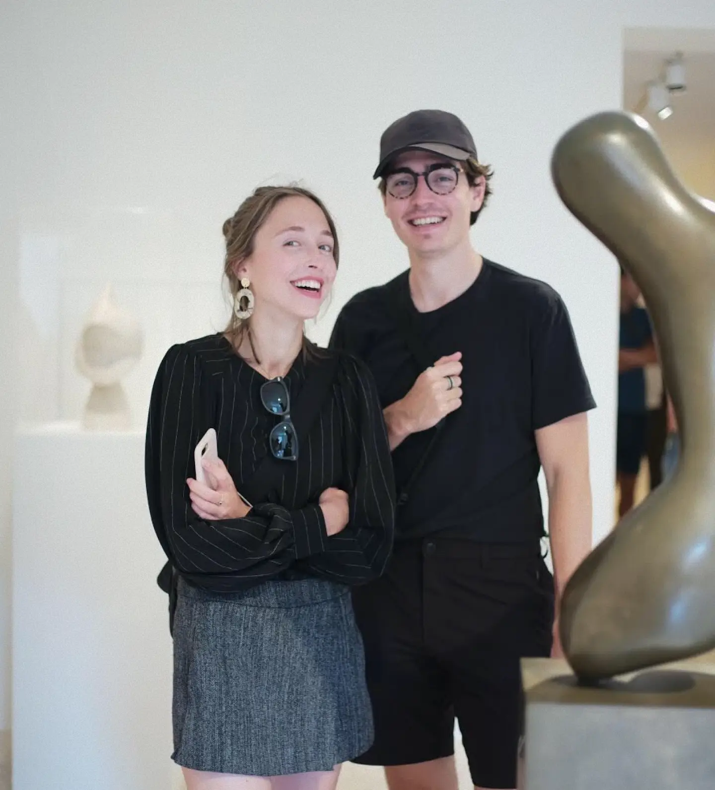 Smiling young woman in a skirt and black blouse standing next to a young man in black shorts and a cap in an art gallery.