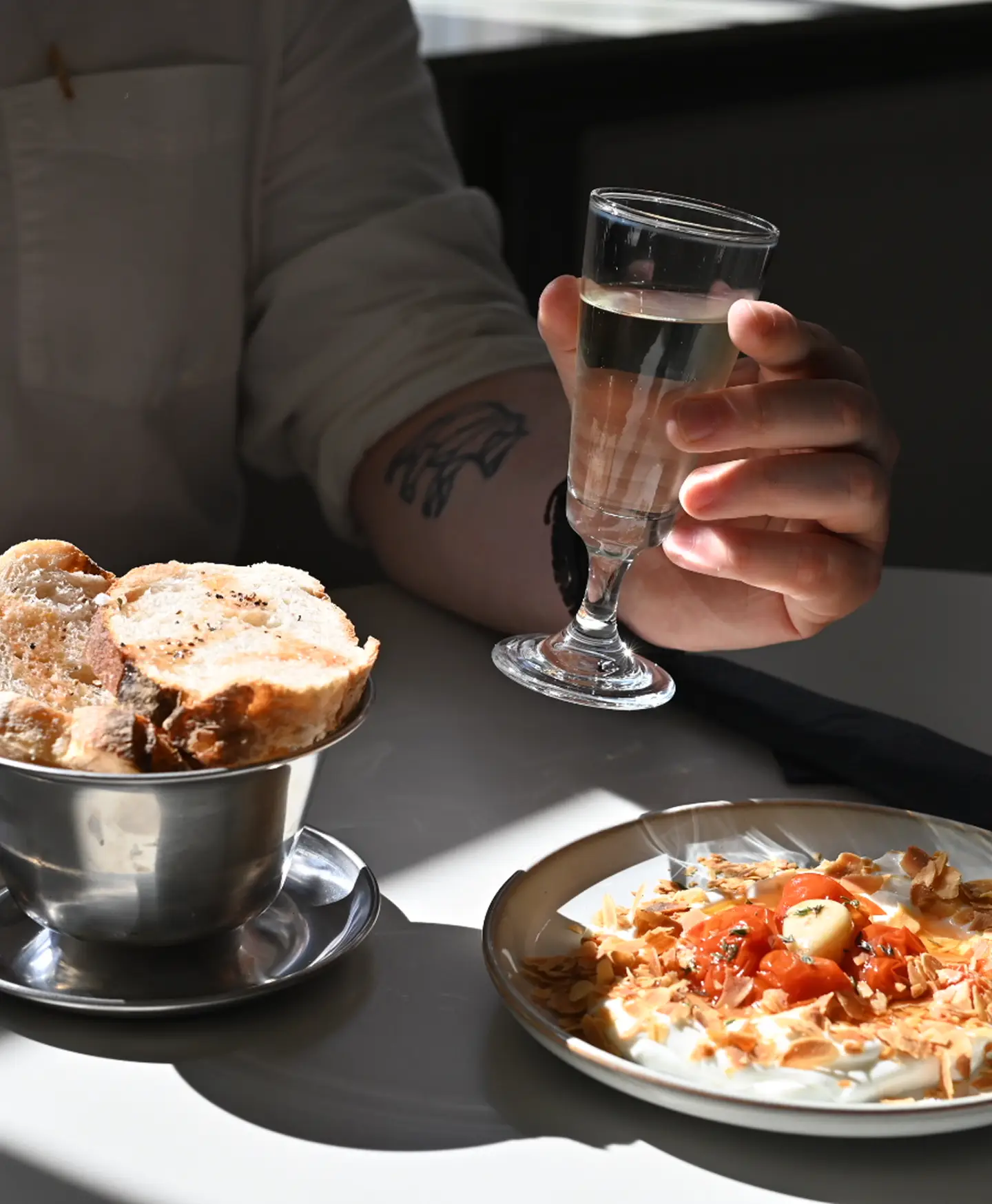 A hand holding a small glass of clear liquid next to a bowl of sliced bread and a plate of food garnished with toasted almonds and cherry tomatoes.