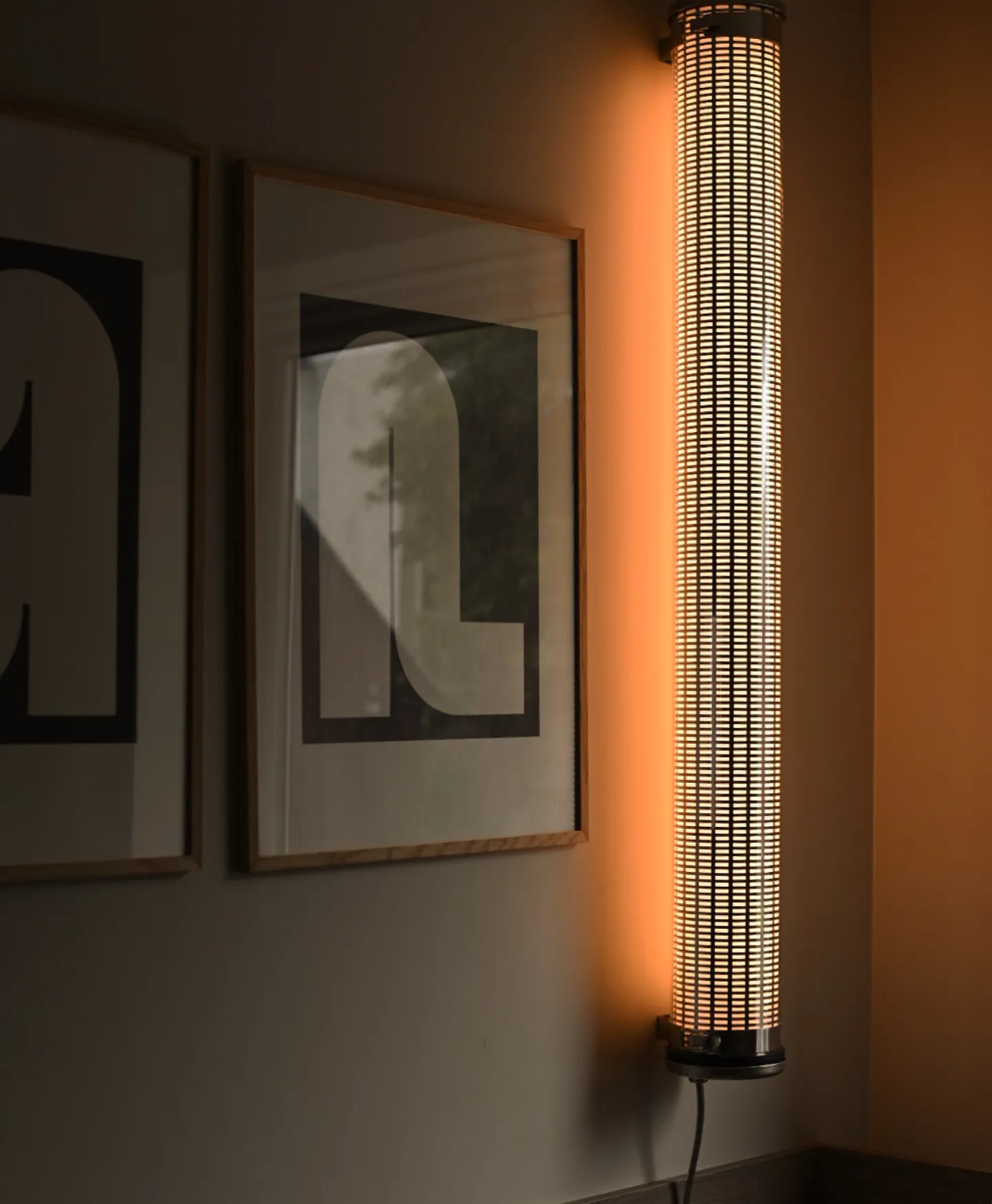 Modern vertical wall lamp emitting warm light next to framed abstract artwork on a wall.