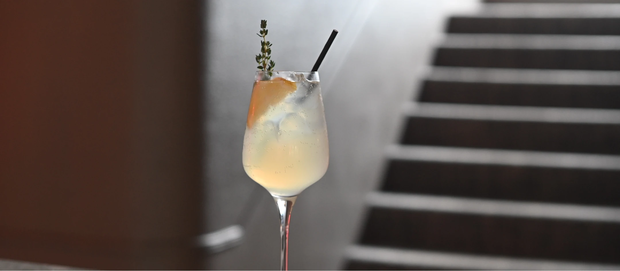 Wine glass with a light yellow cocktail, ice cubes, a lemon slice, a sprig of thyme, and a black straw.