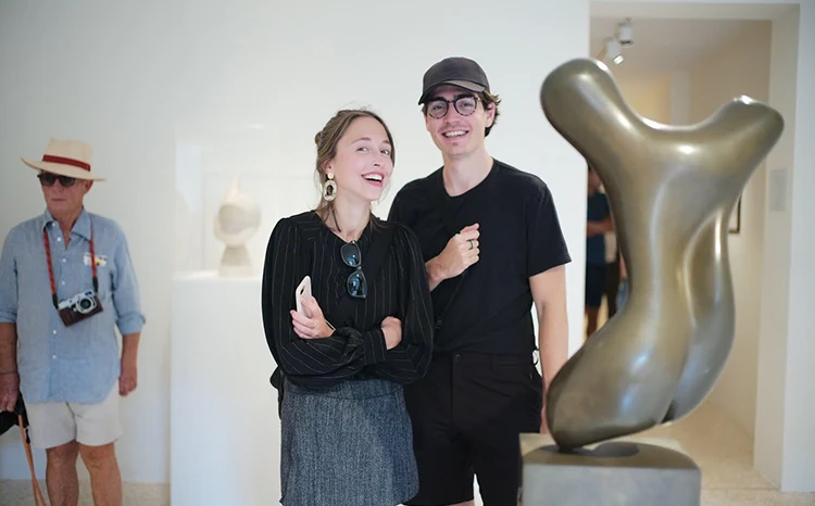 A smiling young couple stands together in an art gallery near an abstract bronze sculpture, with an older man wearing a hat and camera in the background.