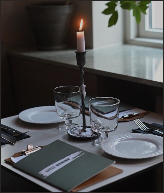 Table set for two with empty white plates, clear glass goblets, cutlery on napkins, a lit candle in a metal holder, and a menu on a clipboard.