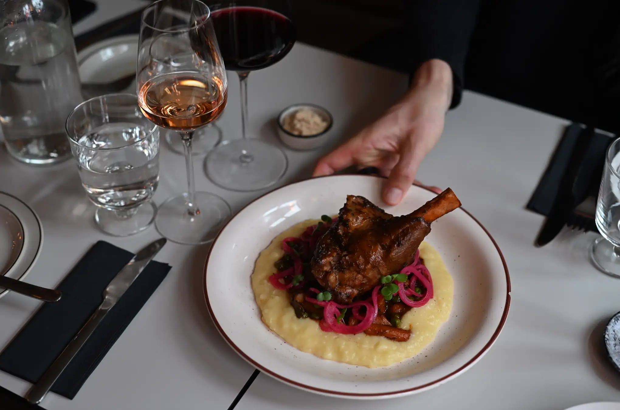 Hand placing a plate with roasted meat on creamy mashed potatoes garnished with pickled red onions and greens, accompanied by glasses of red and rosé wine.
