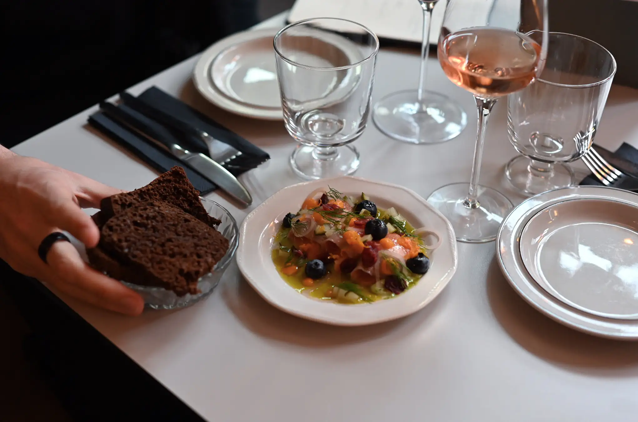 Table set with a plate of gourmet food garnished with blueberries and herbs, a glass of rosé wine, empty glasses, stacked white plates, and a hand holding a bowl of dark rye bread.