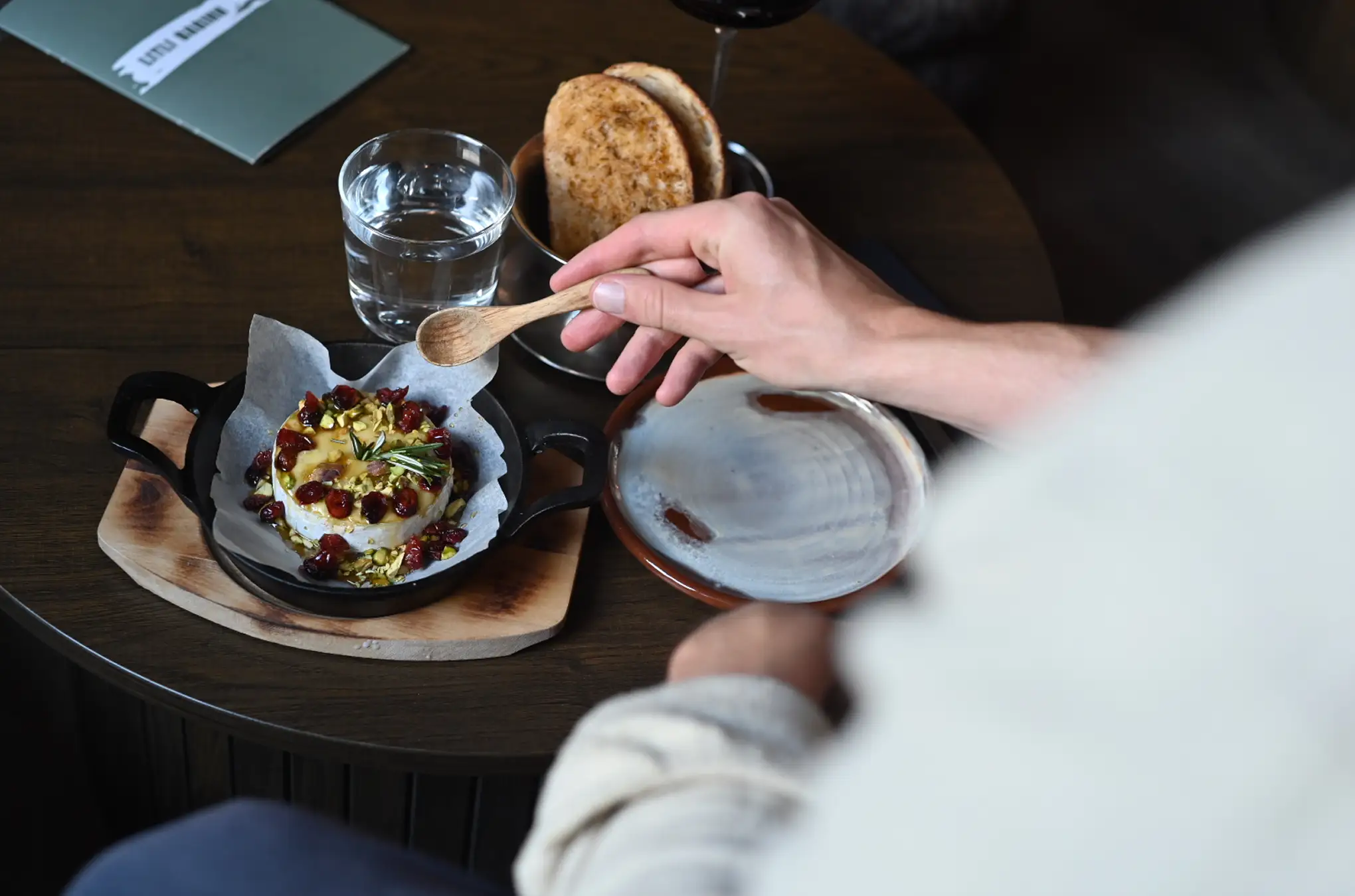 Person holding a small wooden spoon above a round cheese topped with nuts and dried cranberries on parchment paper in a black cast iron dish on a wooden table.