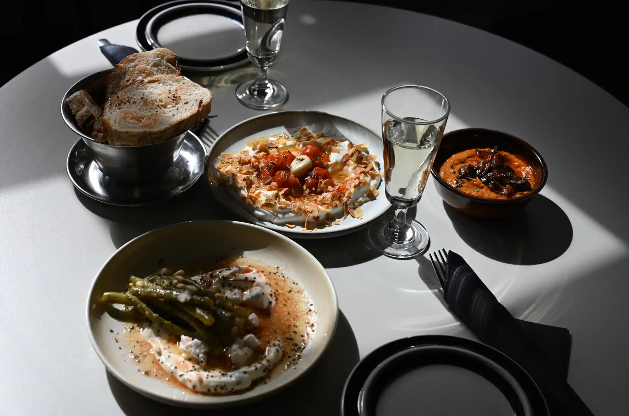 Table set with bowls of assorted dips, toasted bread in a metal cup, two glasses of white wine, and empty black-rimmed plates.