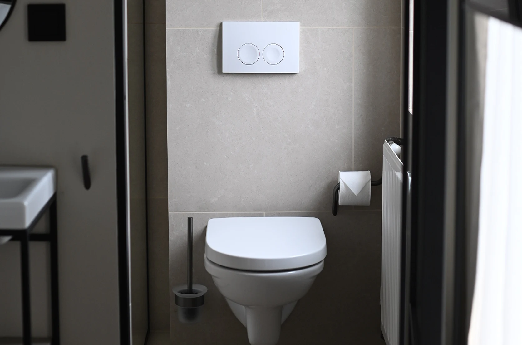 Modern bathroom with a wall-mounted toilet, dual flush buttons, toilet brush, and toilet paper holder on a beige tiled wall.