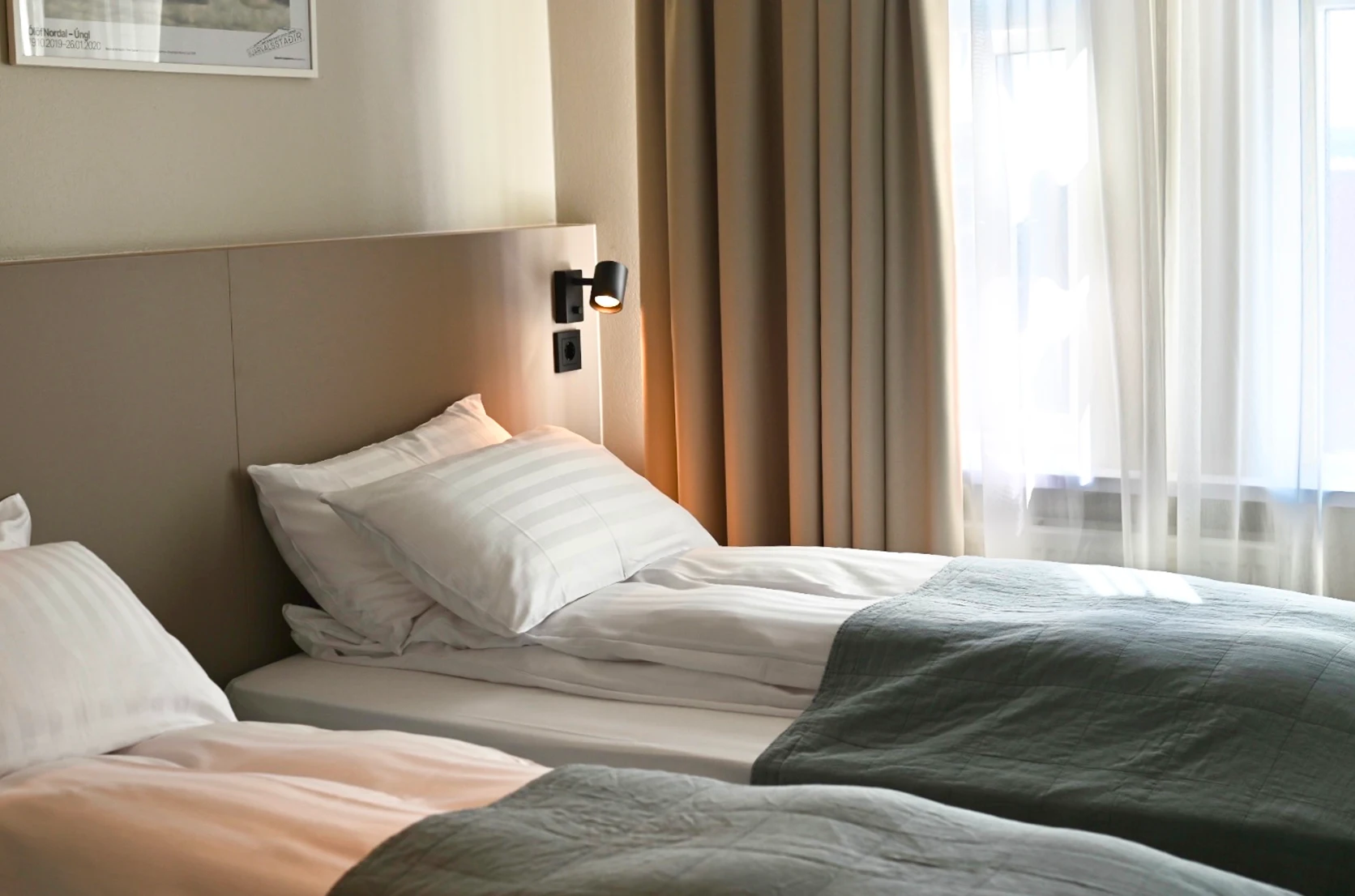 Two neatly made single beds with white striped pillows and grayish-blue quilts beside a window with beige curtains and sheer white drapes.