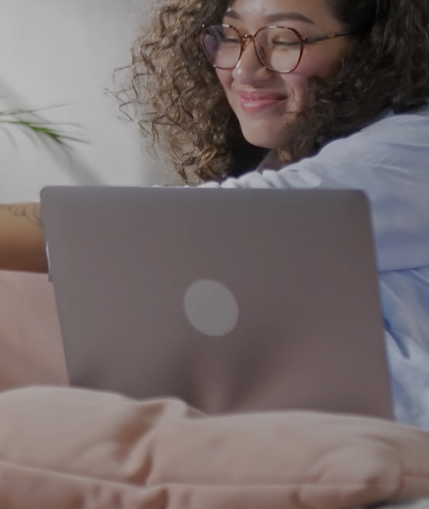 Smiling woman with curly hair and glasses using a laptop while sitting on a couch.