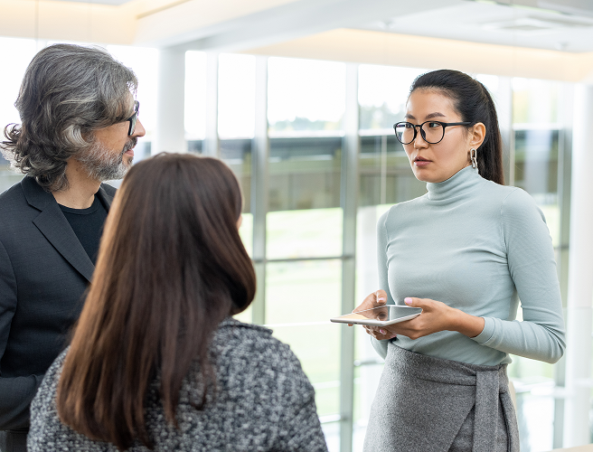Three colleagues engaged in a discussion in a modern office, with one woman holding a tablet.