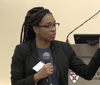 Woman with braided hair and glasses speaking into a microphone while gesturing with her hand.