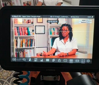 Camera monitor showing a woman in a white shirt sitting at a desk in front of bookshelves with blinds in the background.