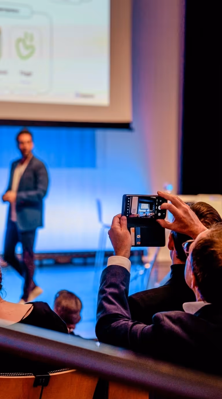 Person filmt einen Mann, der auf einer Bühne vor einer Präsentationsleinwand spricht.