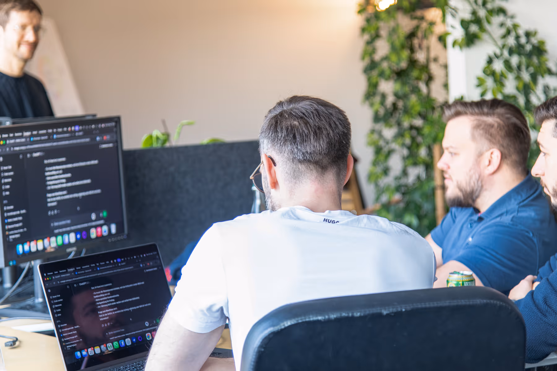 Drei Männer sitzen vor Bildschirmen und diskutieren über Programmiercode in einem modernen Büro mit Pflanzen im Hintergrund.