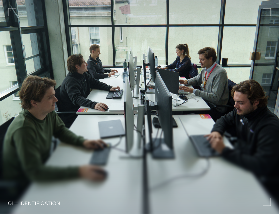 Six people working at computers arranged in two rows facing each other in an office with large windows.