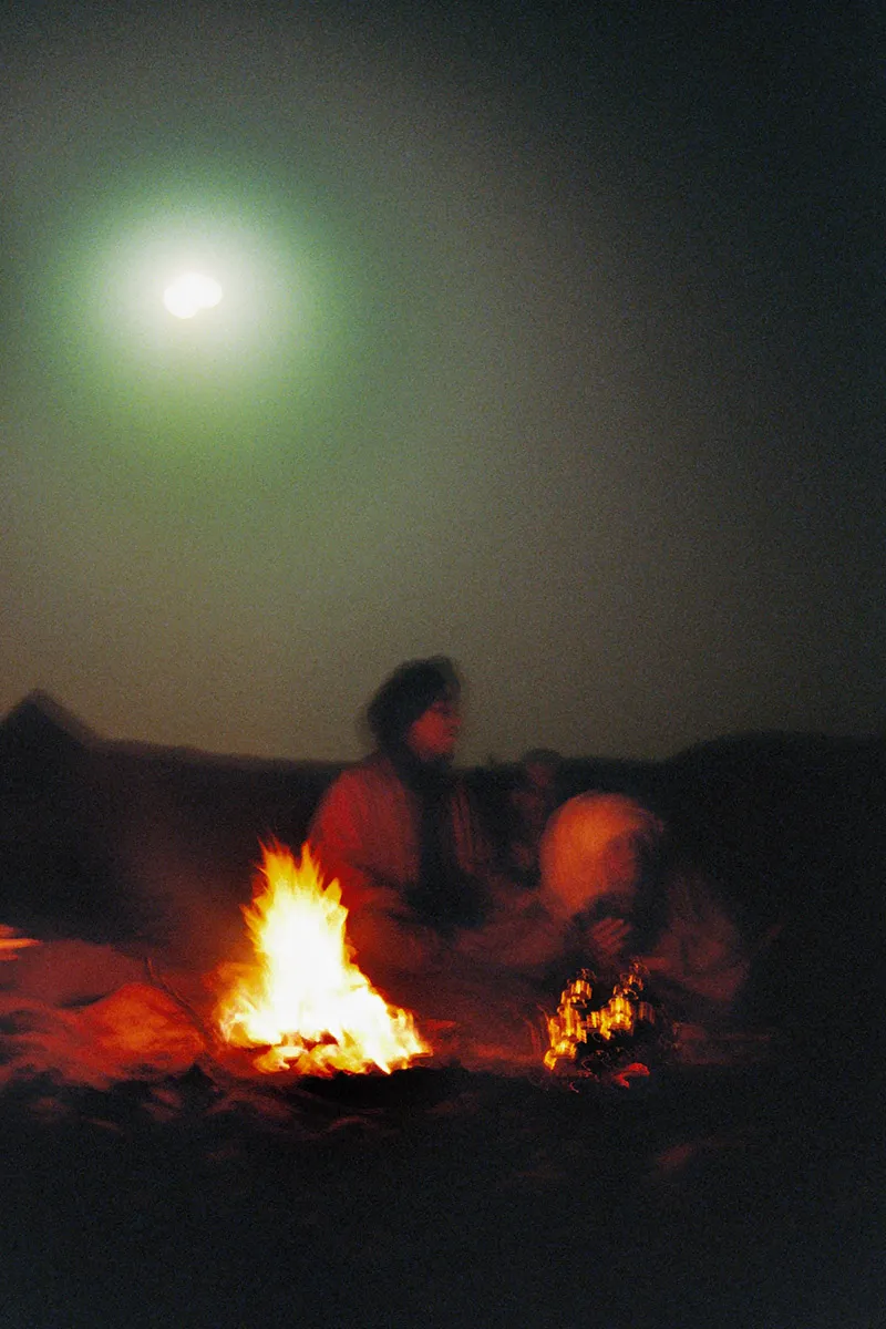 Two men sitting around a camp fire in the desert with the moon illuminating the night sky.