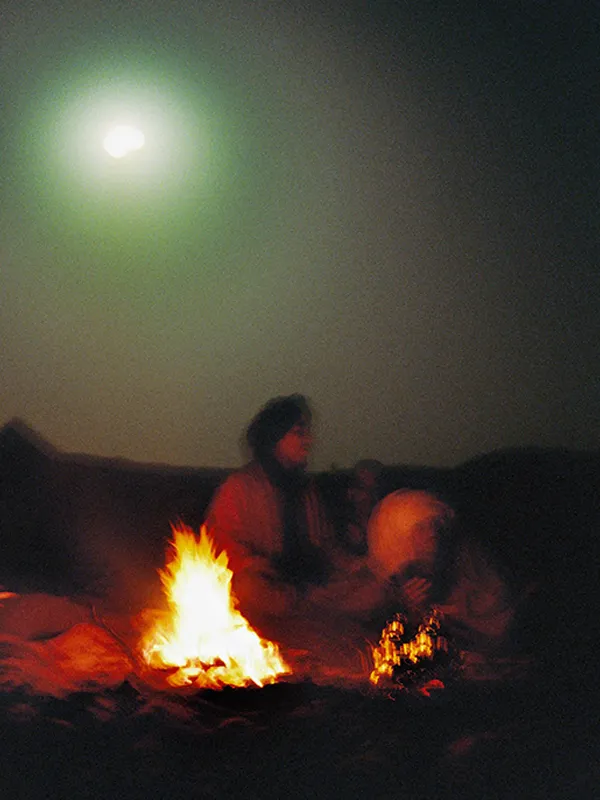 Two men sitting around a camp fire in the desert with the moon illuminating the night sky.