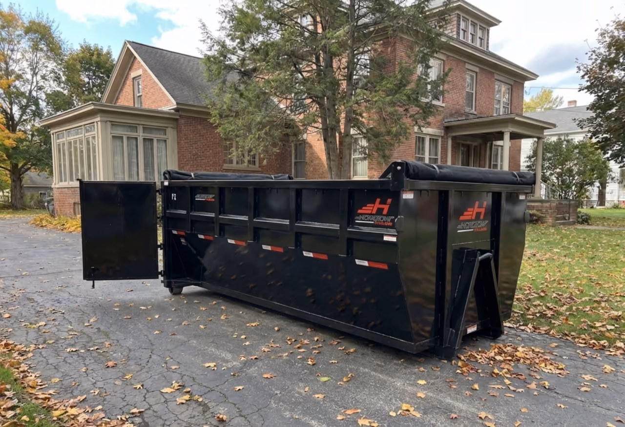 Large black industrial dumpster with door open on cracked driveway near a brick house with trees.