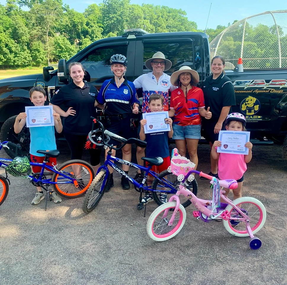 Group of children holding certificates and standing with adults, bicycles, and a black truck with D.A.R.E. logos in the background on a sunny day.
