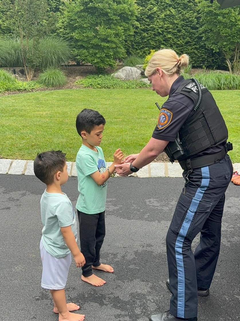 Police officer handcuffing a young boy while another barefoot boy watches on a driveway with green lawn and shrubs in the background.