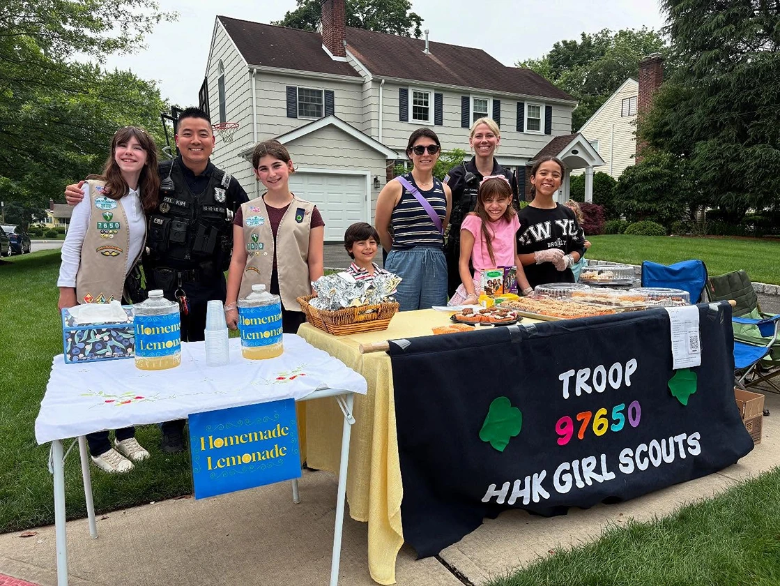 Group of Girl Scouts and two police officers standing behind a table with homemade lemonade and baked goods outside a suburban house.
