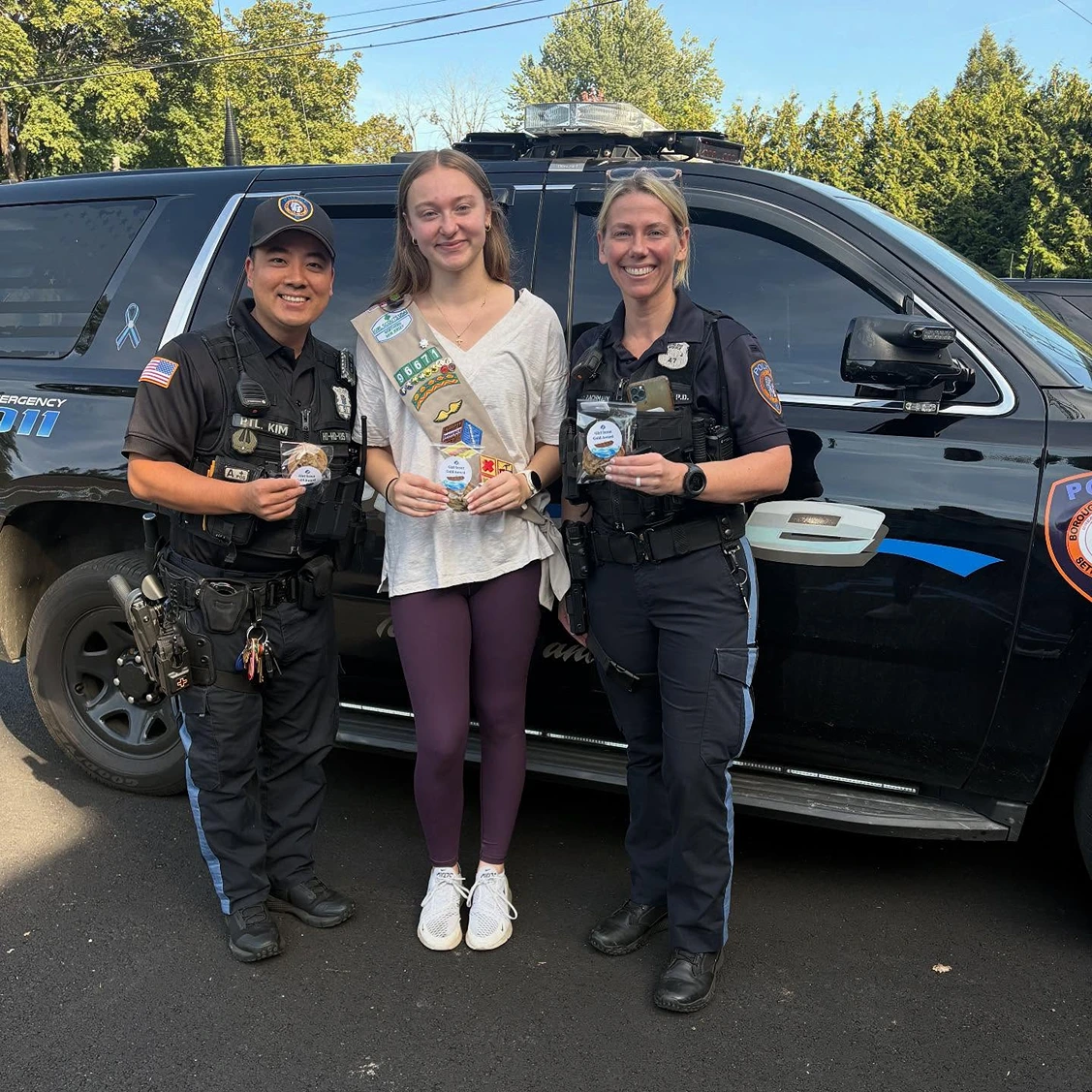 Two police officers and a Girl Scout standing in front of a police SUV, all smiling and holding small packaged items.