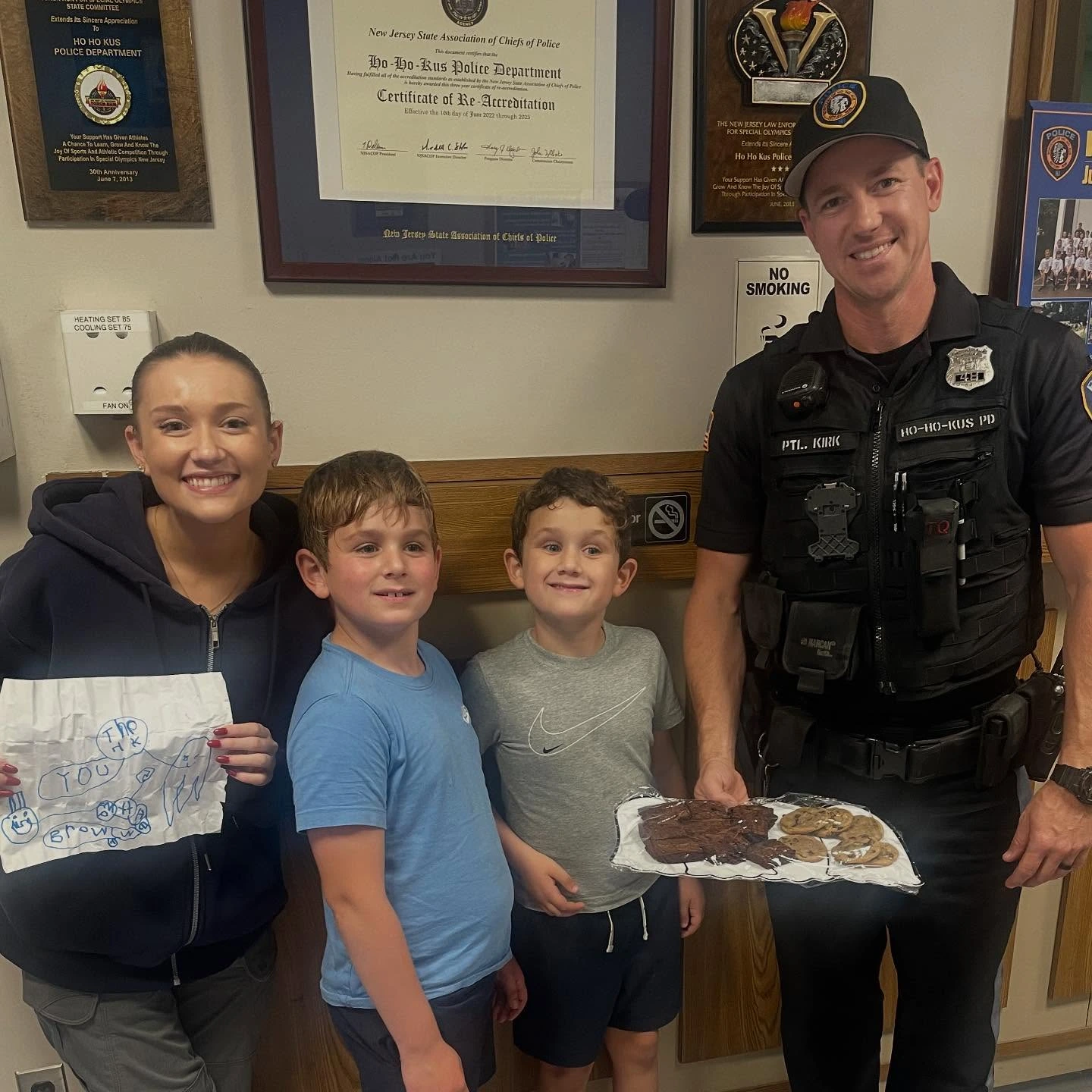 Smiling woman holding a thank-you note stands next to two boys and a smiling police officer holding a tray of cookies.