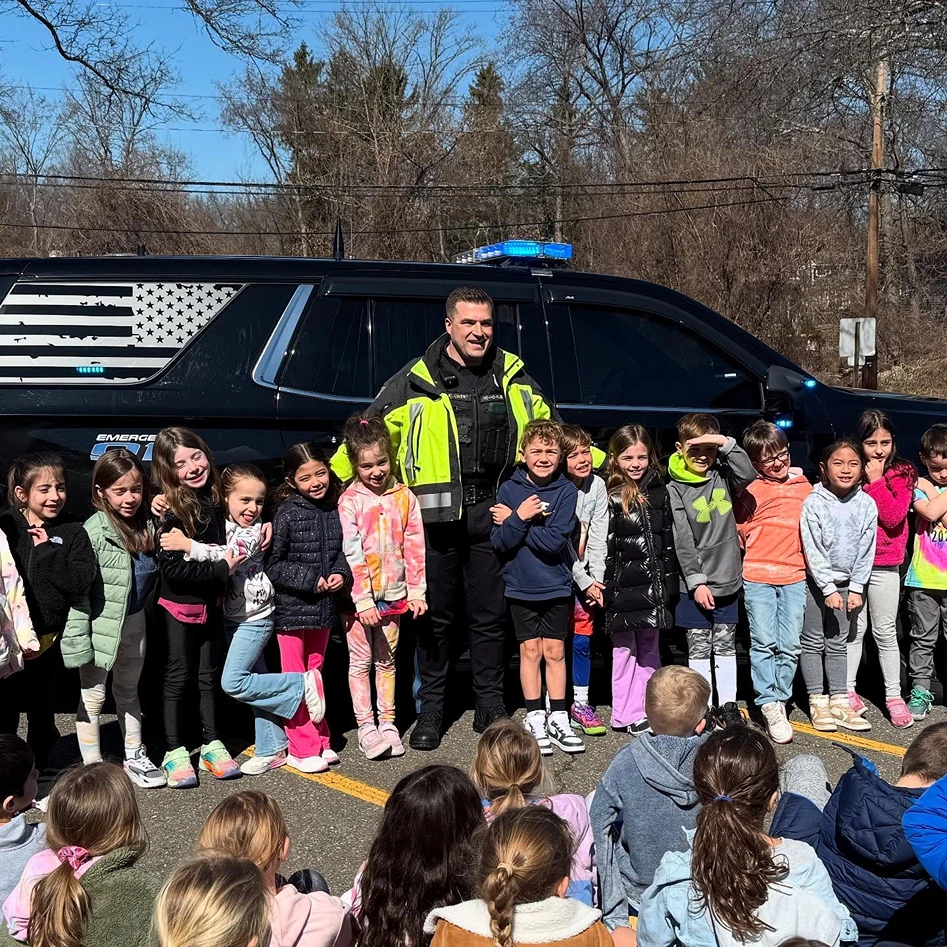 Police officer in a bright jacket posing and smiling with a group of children standing in front of a police SUV with an American flag design on the window.