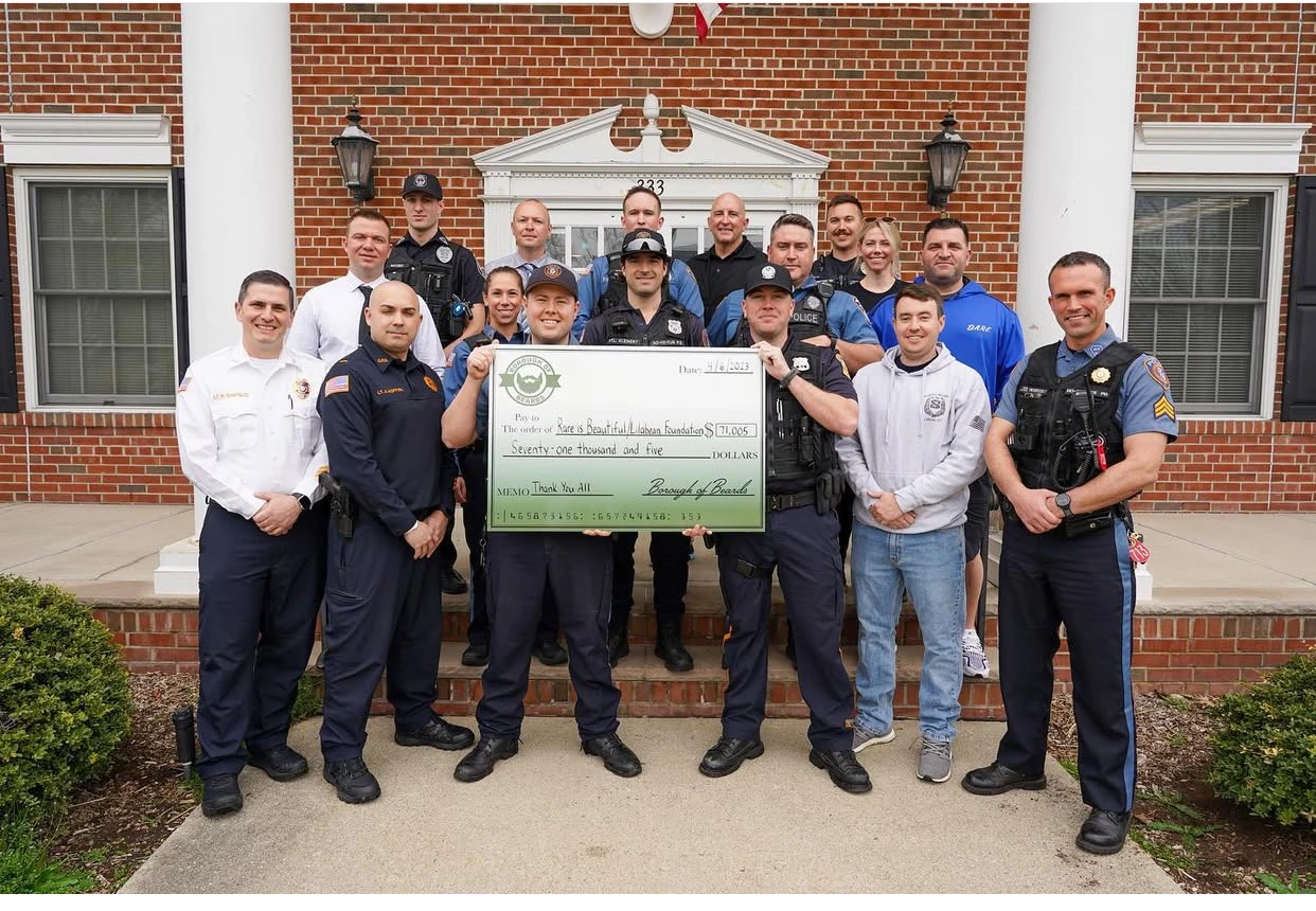 Group of uniformed police and emergency personnel standing in front of a brick building holding a large donation check for $71,005 to the Rare is Beautiful/Hiabean Foundation.