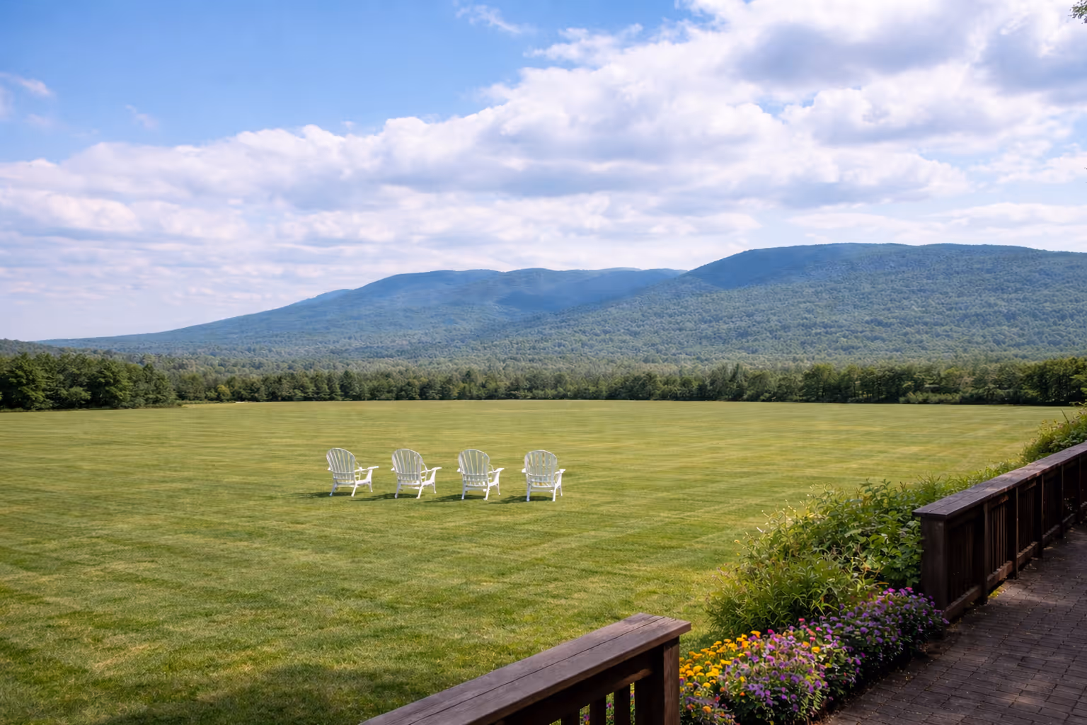 Four white Adirondack chairs on a large green lawn facing forested mountains under a partly cloudy sky.