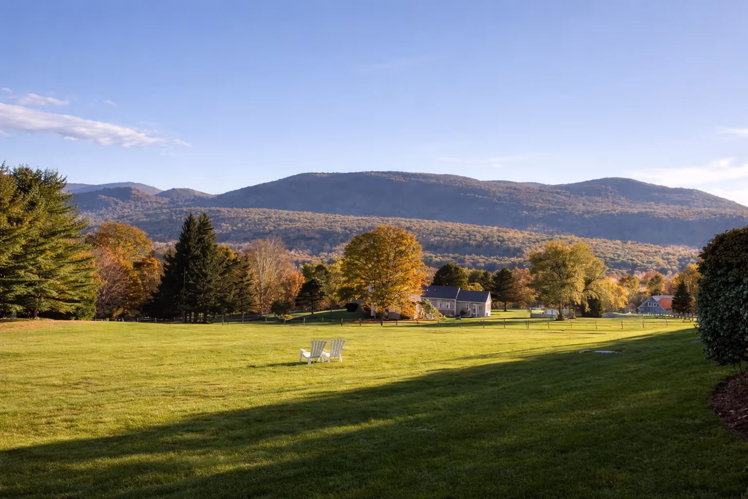 Sunlit green lawn with two white chairs, colorful autumn trees, houses, and forested mountains under a clear blue sky.