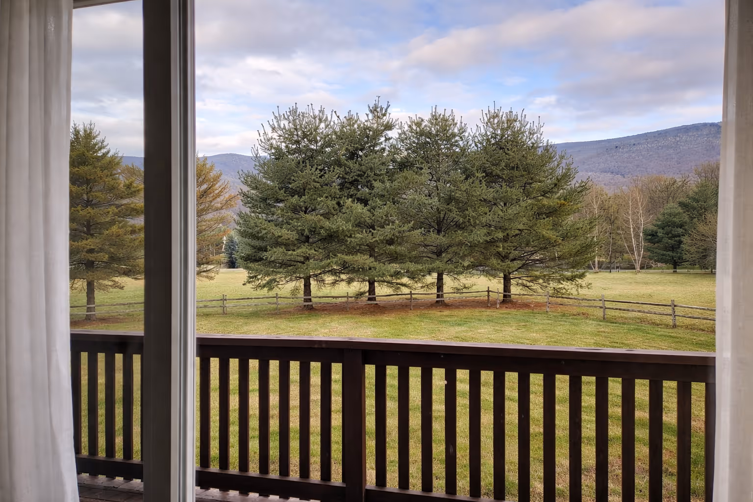 View through a window and balcony railing of green pine trees, grassy field, wooden fence, and distant mountains under a partly cloudy sky.