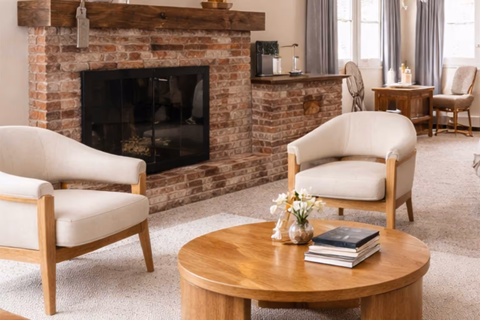 Cozy living room with beige armchairs, round wooden coffee table with books and flowers, and a brick fireplace with wooden mantle.