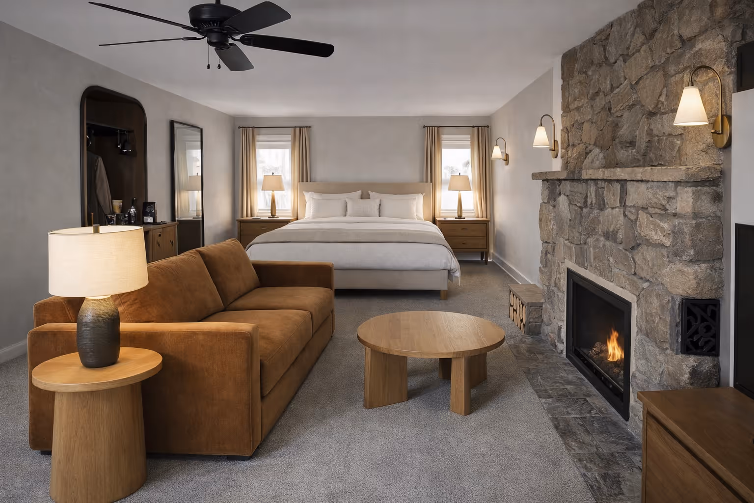 A modern bedroom with a brown sofa, round wooden coffee table, stone fireplace, and a bed between two windows with beige curtains and lamps.