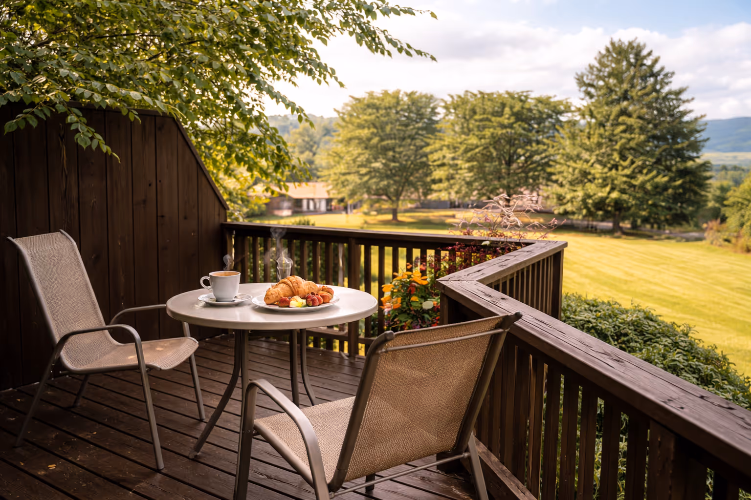 Private wooden terrace with two chairs and a round table holding a steaming cup of coffee and a plate with croissants and fruit, overlooking a green landscape with trees and a house in the distance.