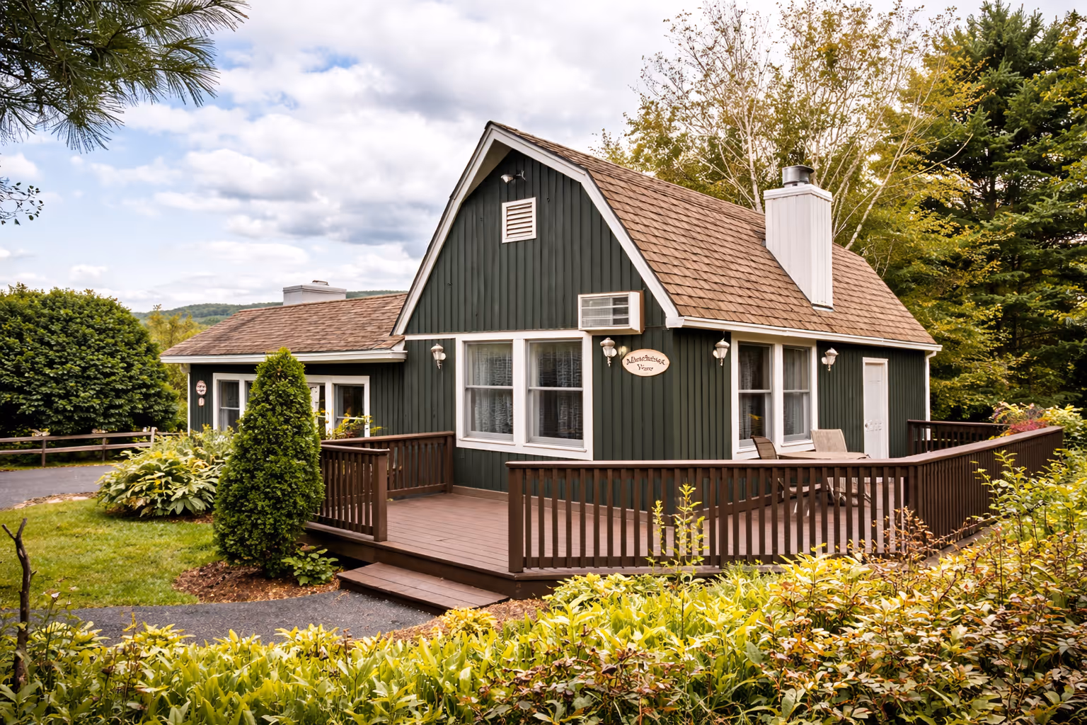 Green wooden cottage with brown roof and wraparound deck surrounded by lush green plants and trees.
