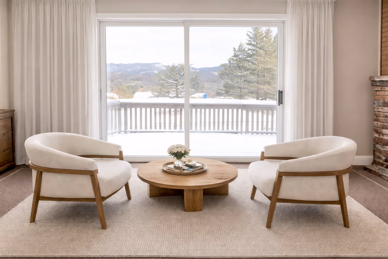Cozy sitting area with two white cushioned chairs, a round wooden coffee table with flowers, and a large sliding glass door overlooking a snowy terrace and pine trees.