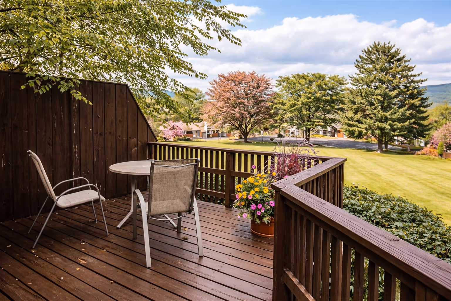 Wooden terrace with two chairs, a round table, and a flower pot overlooking a green lawn and trees under a partly cloudy sky.