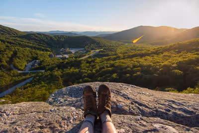 View from a rocky hill with hiking boots visible, overlooking a green valley and hills under a sunrise or sunset sky.