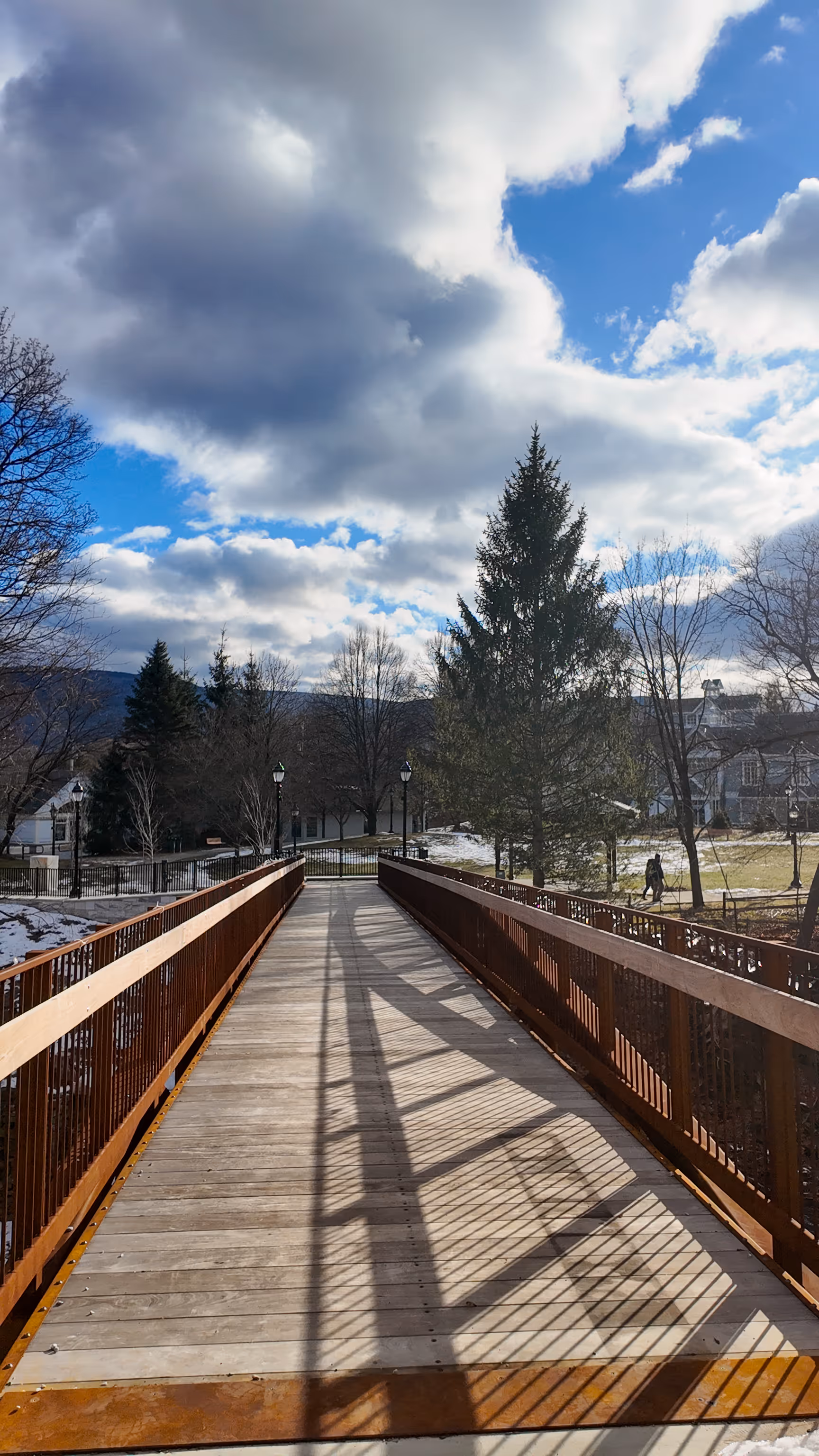 Wooden pedestrian bridge with metal railing casting shadows, leading to a park with trees and cloudy blue sky.