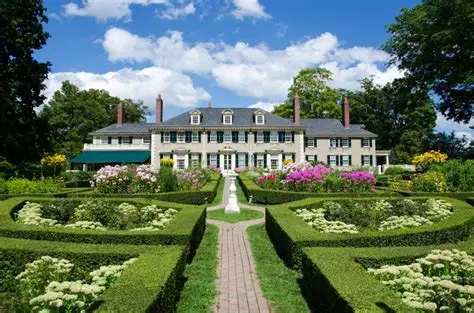 Large white colonial-style mansion with symmetrical windows, surrounded by manicured gardens and hedges under a blue sky with clouds.