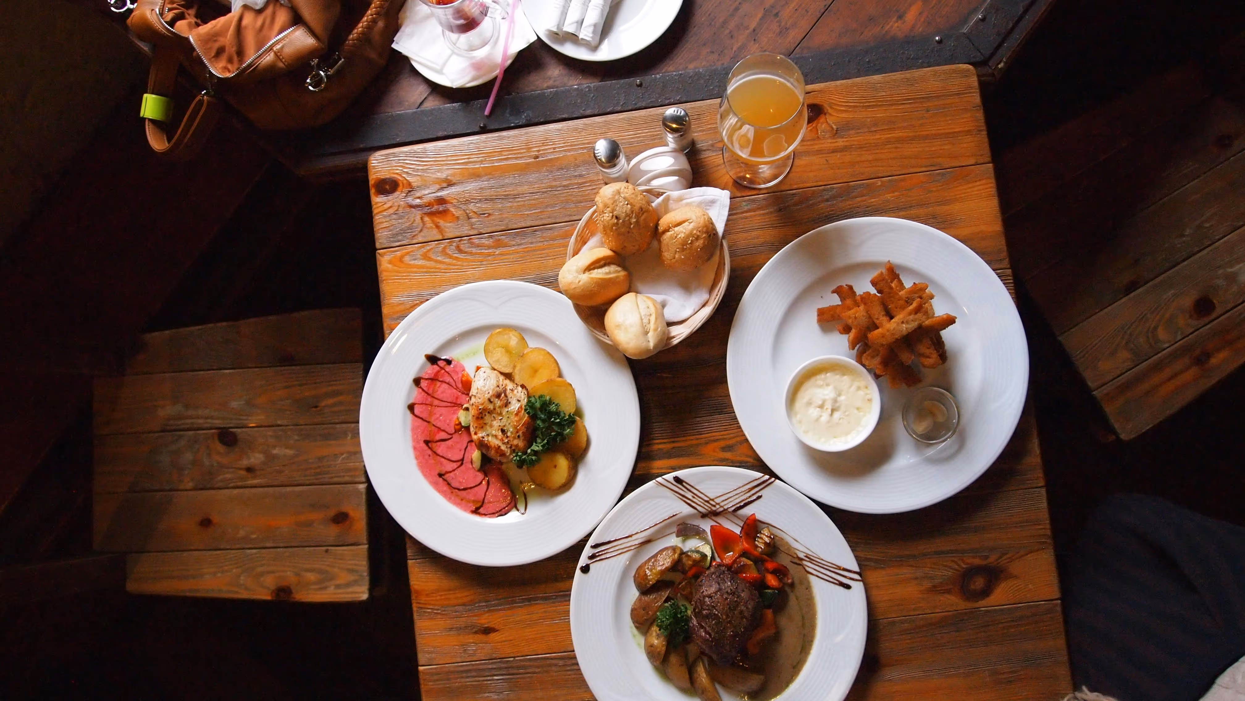Rustic wooden table with three plates of restaurant dishes including a grilled chicken salad, steak with vegetables and potatoes, and fried sticks with dipping sauce, alongside a basket of bread rolls and a glass of drink.